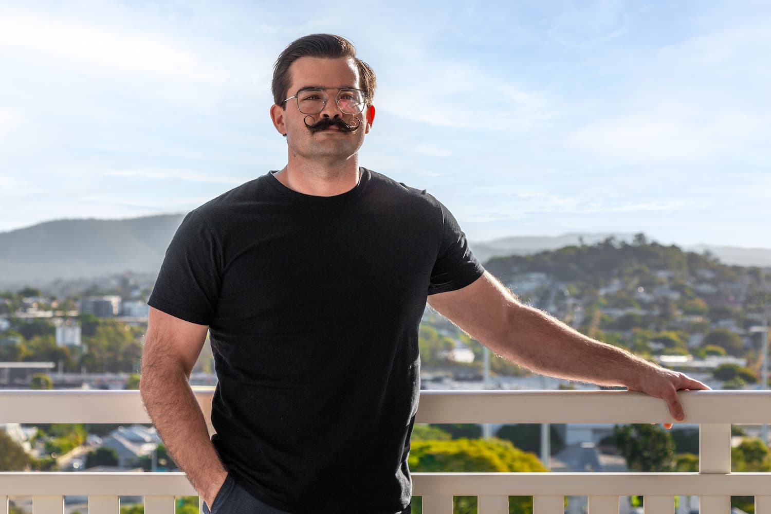 A brunette man in a black shirt with a moustache and glasses, posing for the camera